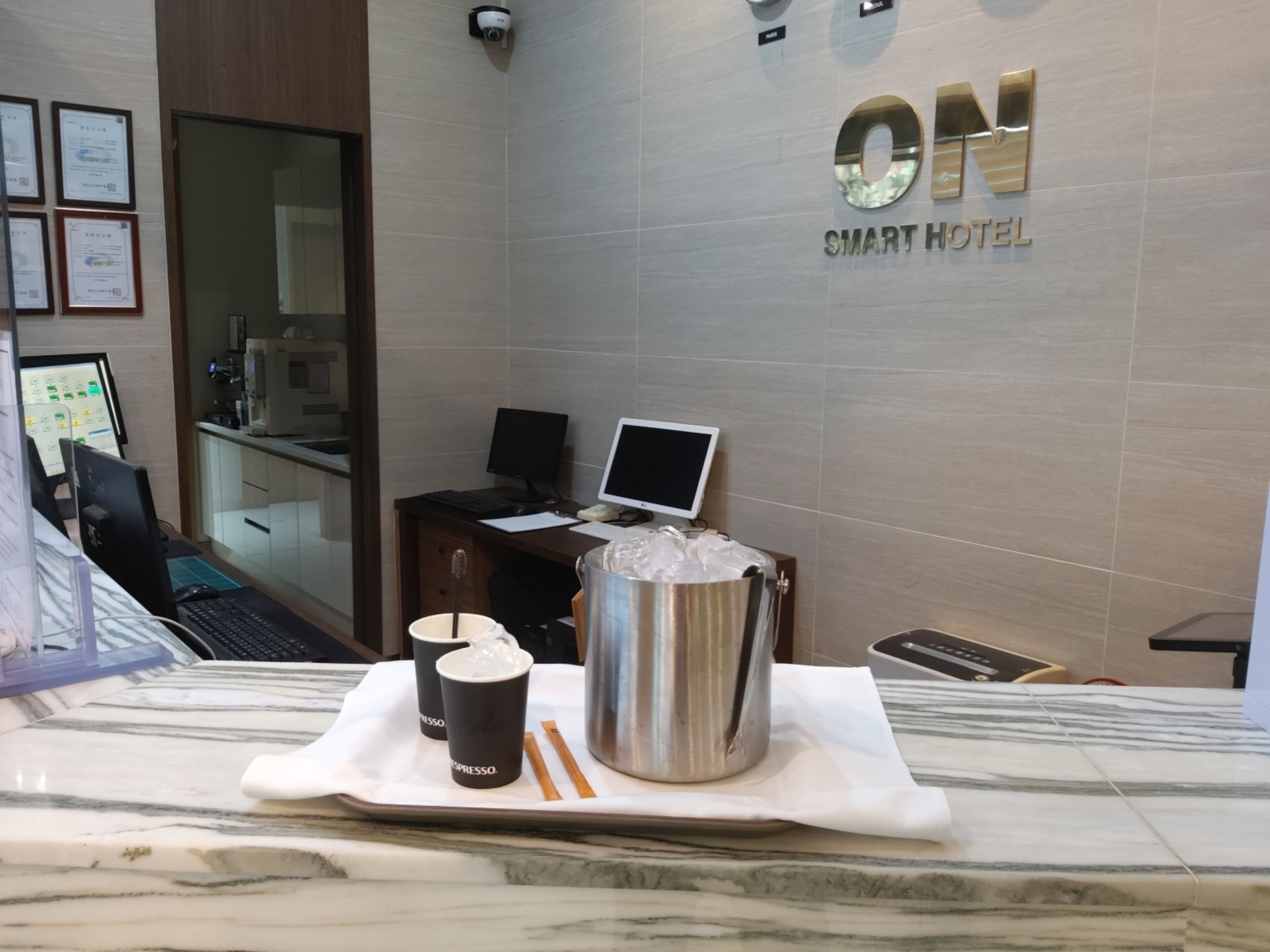 Zona de recepción de un hotel con mostrador, computadora y estación de café con dos espressos y una termo.