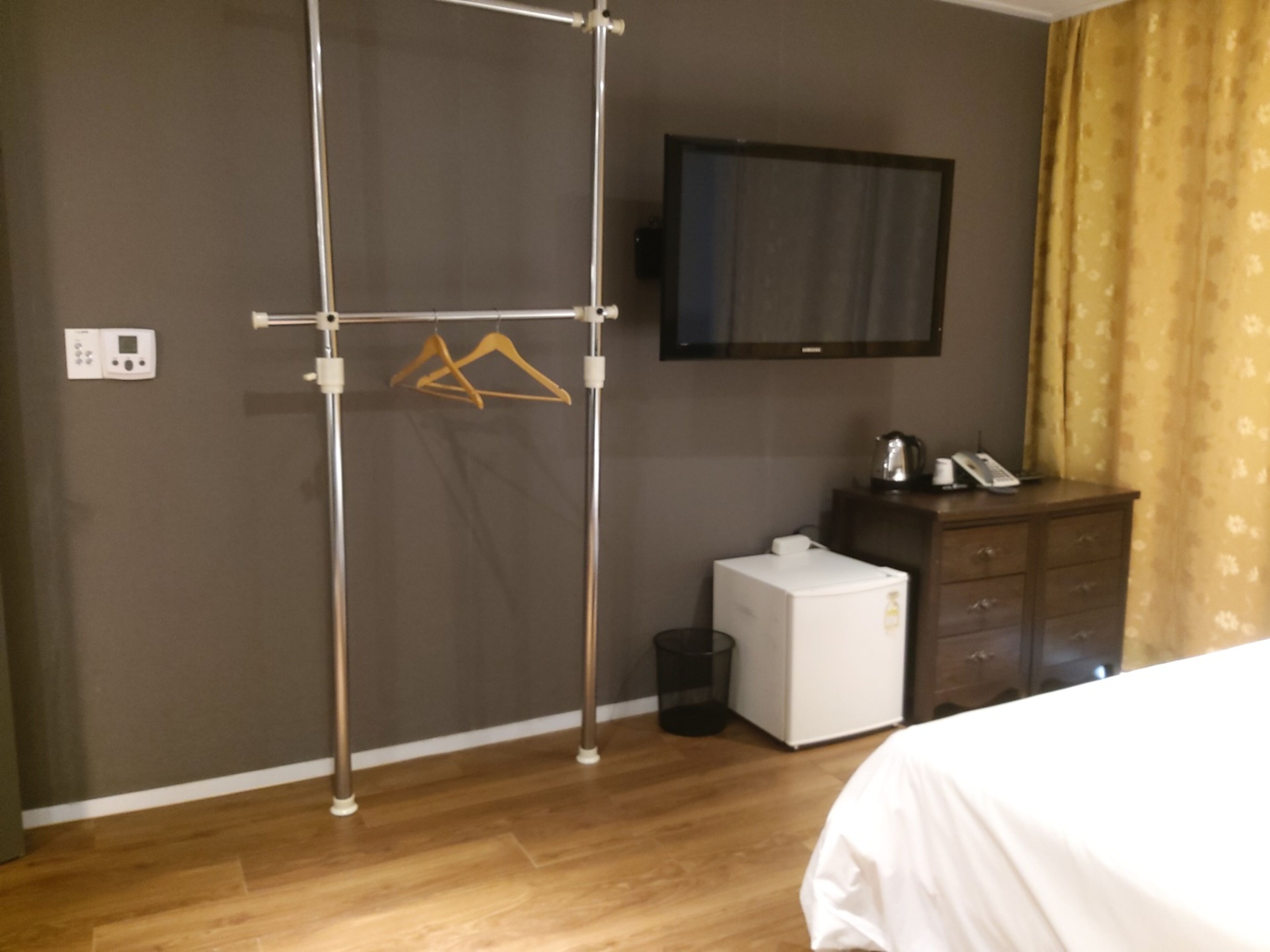 A hotel room with a wooden floor, a bed, a small white refrigerator, a wooden dresser, a television mounted on the wall, and a metal rack with hangers.