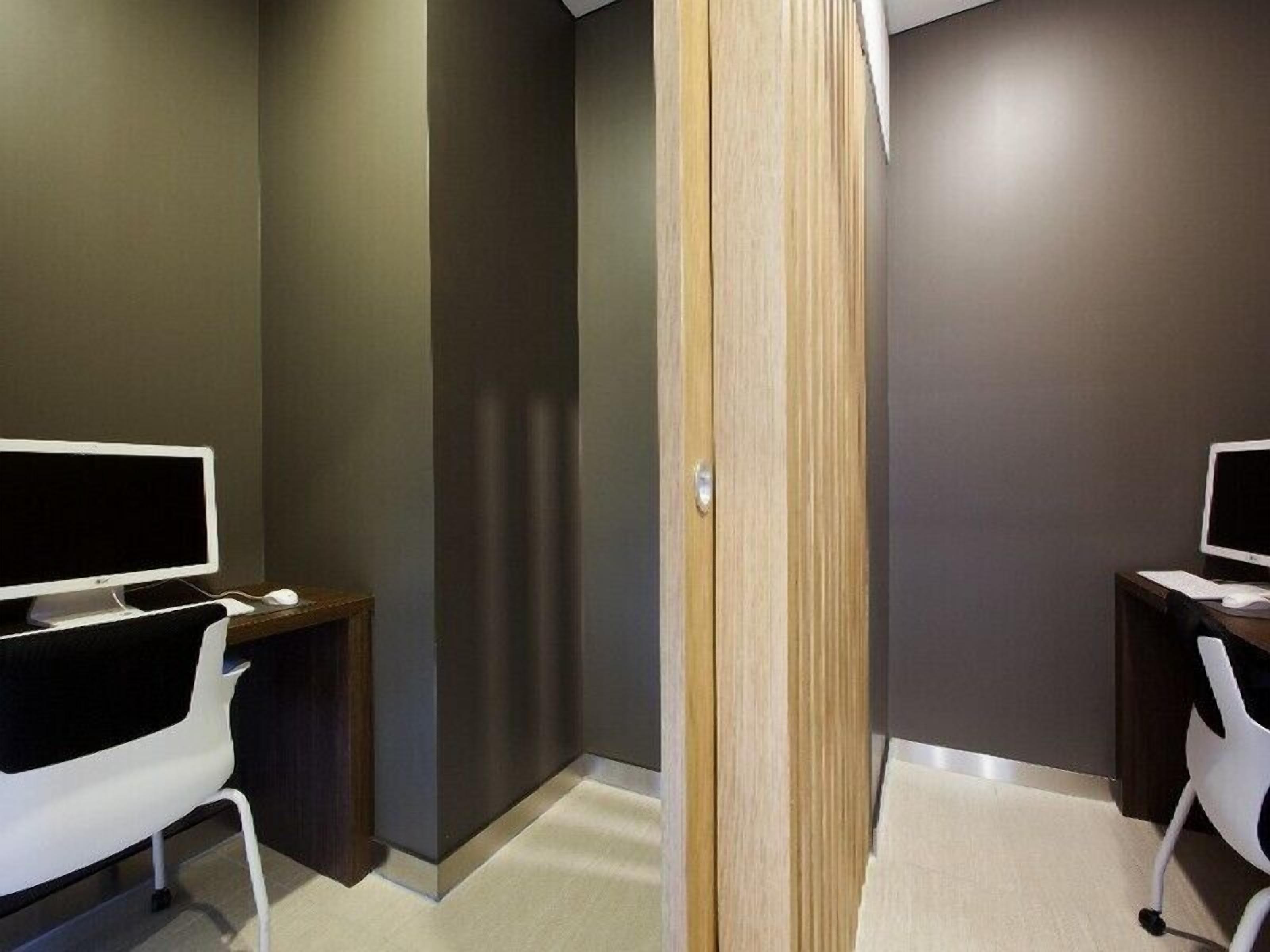 Un espacio de oficina moderno con dos escritorios, cada uno con un monitor de computadora, un teclado y un ratón. La habitación tiene una decoración en tonos neutros, con paredes oscuras y suelo claro.