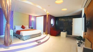 Habitació d'hotel moderna amb un llit corbat, un otomà vermell, una taula auxiliar i un televisor fixat a la paret.