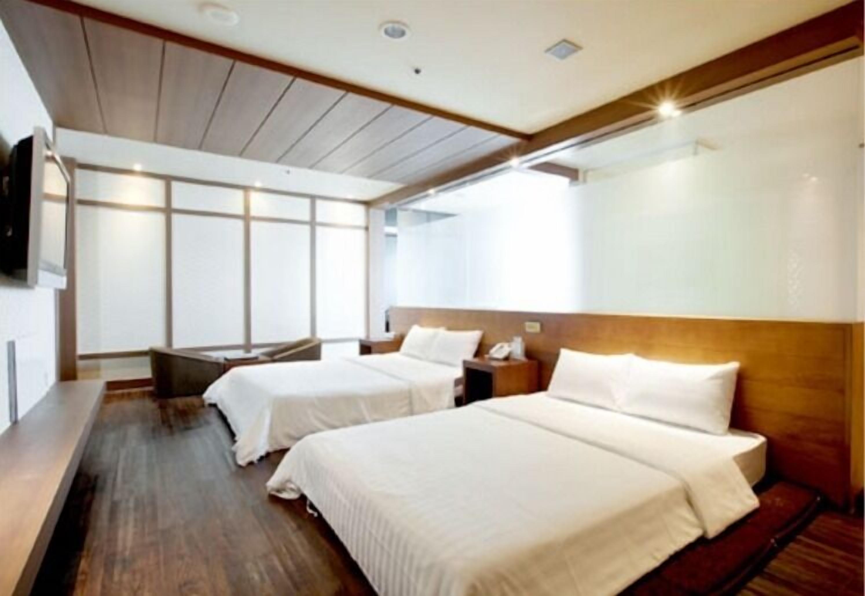 A hotel room with two beds, wooden headboards, a bench, and a large window.