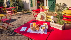 Een terras met rode tafels en stoelen, een mand met brood, een croissant, een kom soep en een glas sinaasappelsap.