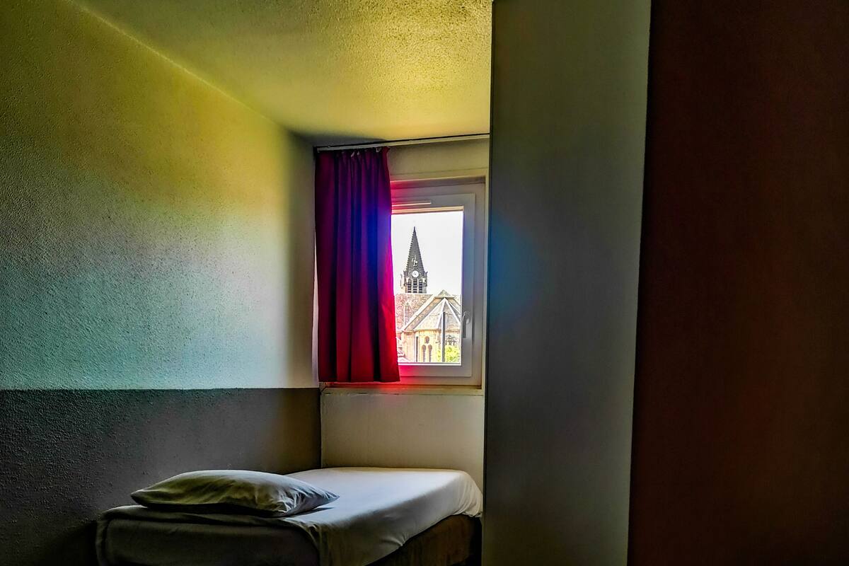 Ein kleiner, schummrig beleuchteter Raum mit einem Bett, einem Fenster mit roten Vorhängen und Blick auf ein Gebäude mit einem Kirchturm.