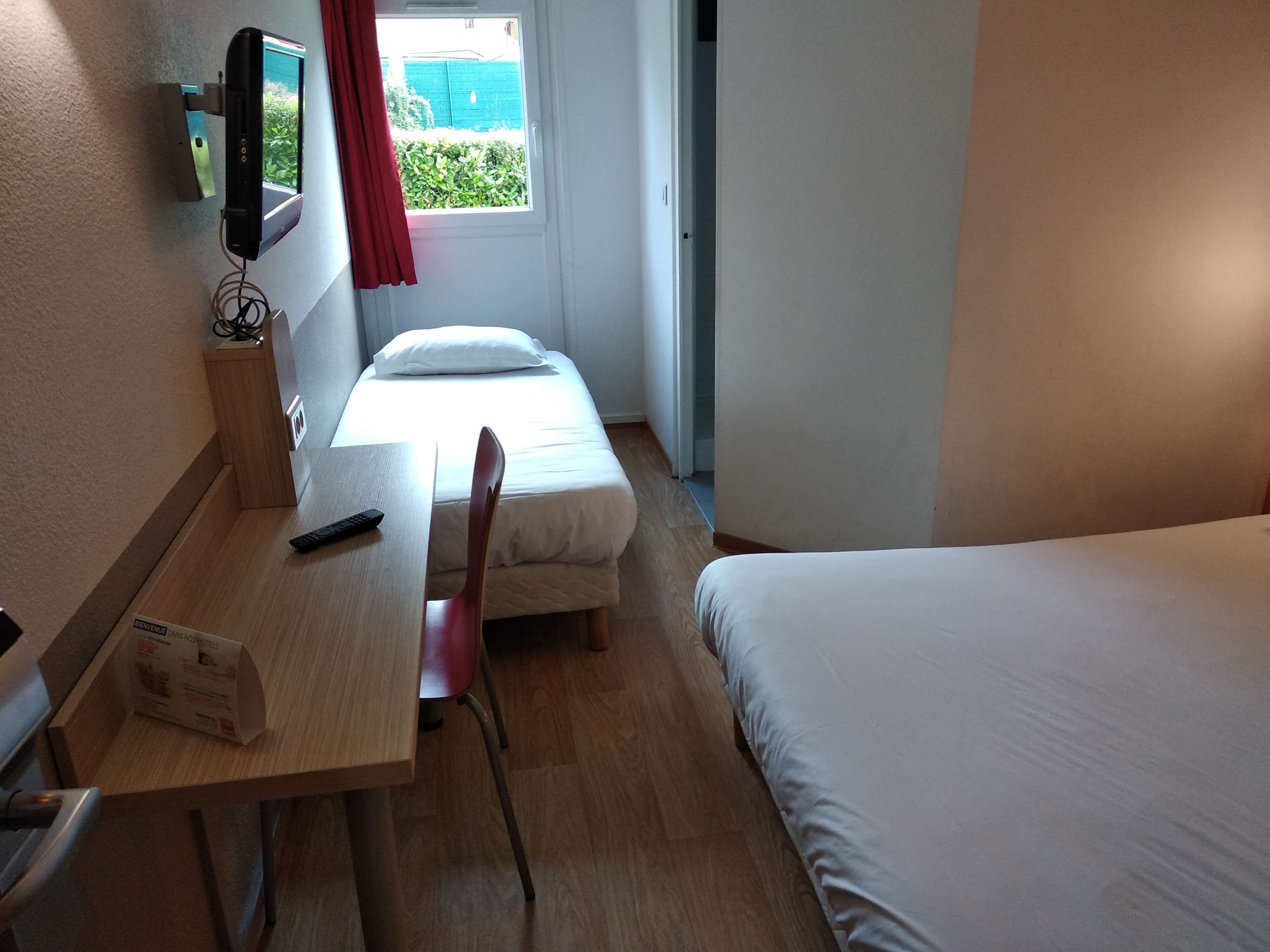 A hotel room with a bed, a desk with a chair, a TV mounted on the wall, and a window with a curtain.