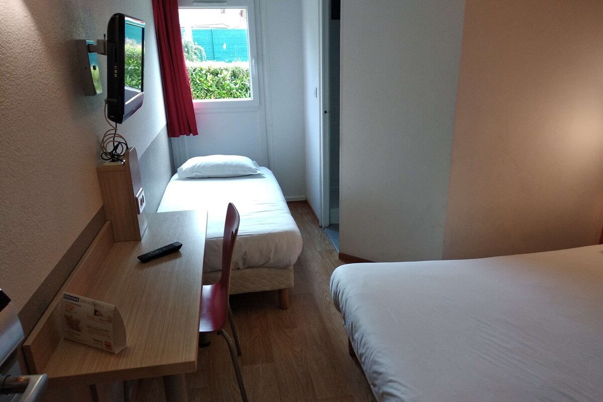 A hotel room with a bed, a desk with a chair, a TV mounted on the wall, and a window with a curtain.