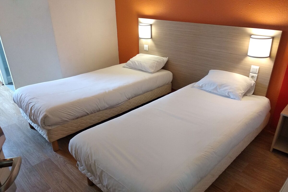 A hotel room with two single beds, each with a pillow and a headboard with a light fixture.