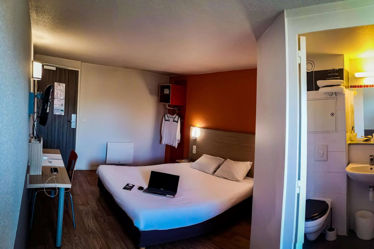Ein Hotelzimmer mit Bett, Schreibtisch mit Laptop, Stuhl, Nachttisch und einem Badezimmer mit Waschbecken und Toilette.