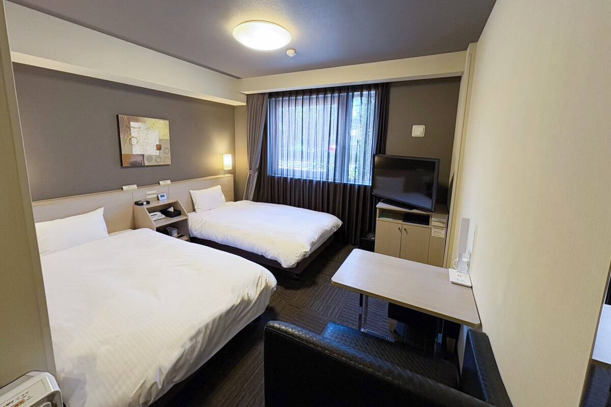 A hotel room with two beds, a desk, a television, and a window with curtains.
