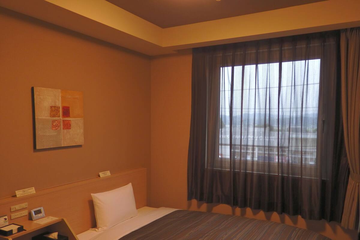 A hotel room with a bed, a desk with a telephone and a clock, a window with curtains, and a wall-mounted artwork.