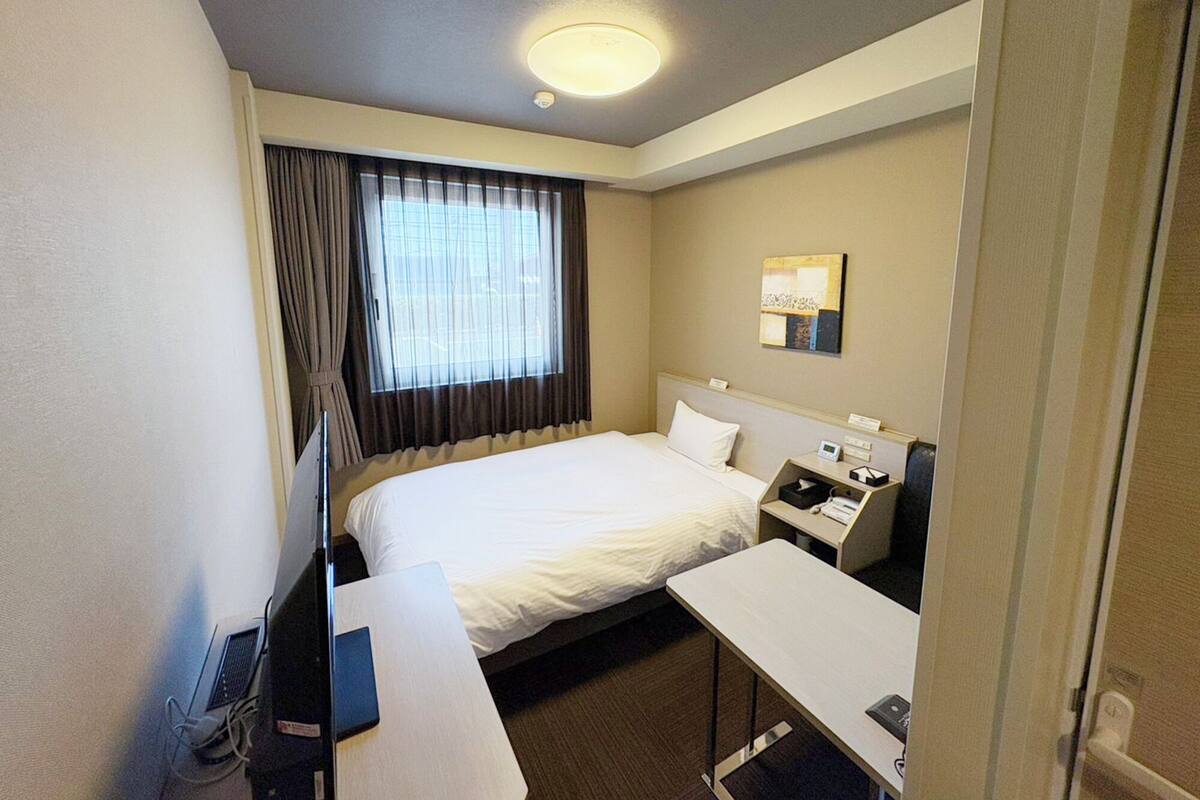 A hotel room with a bed, a desk, a chair, a bedside table, a lamp, a window with curtains, and a wall-mounted light fixture.