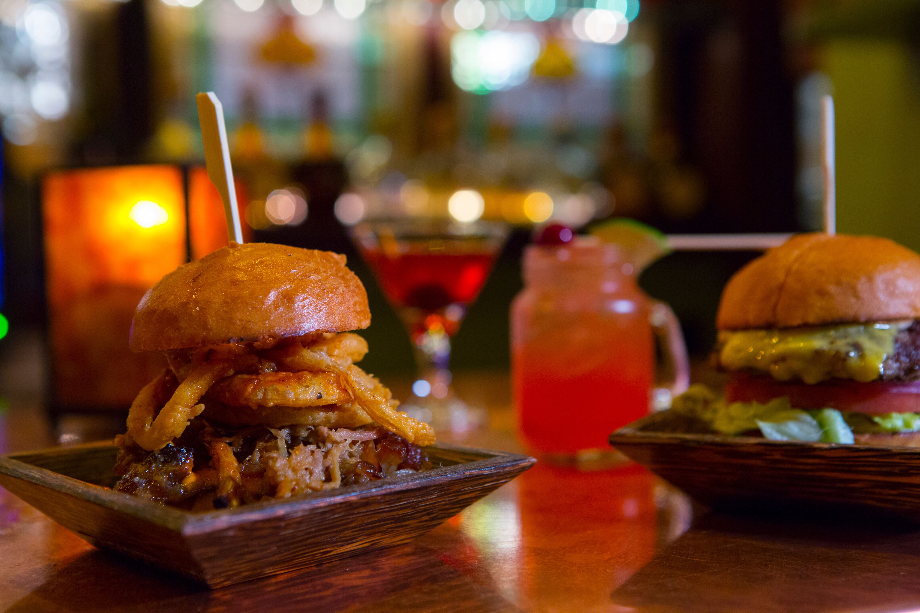 Un bar con dos hamburguesas en platos de madera, un cóctel y una vela encendida.