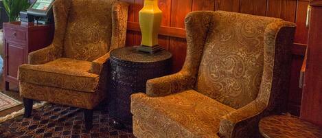 A lobby with two patterned armchairs, a small round table, a large ornate mirror, and a potted plant.