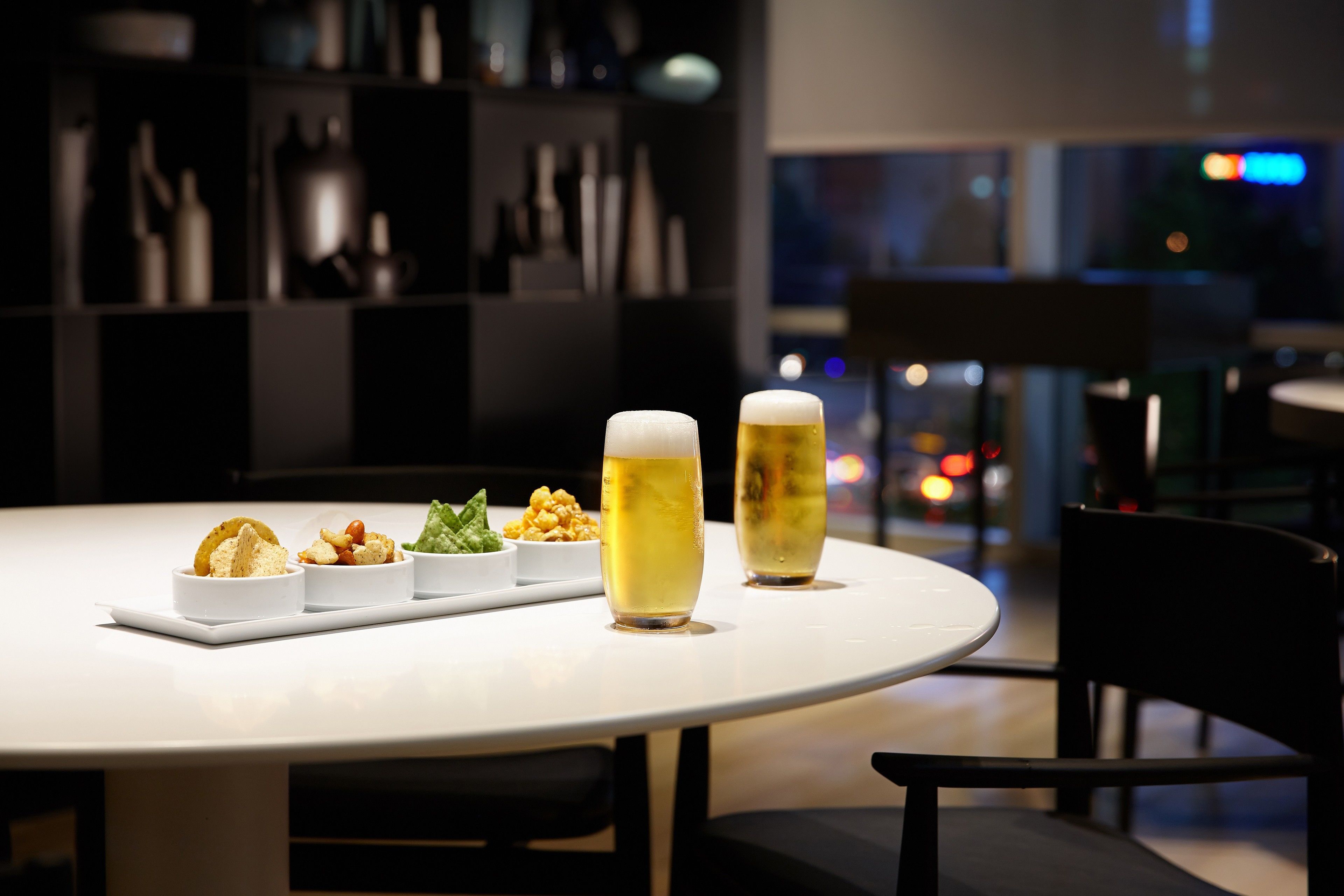 Una mesa redonda con superficie blanca, dos vasos de cerveza y tres platos pequeños de comida.