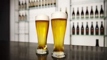 Dos vasos altos de cerveza sobre una barra con estantes llenos de botellas al fondo.