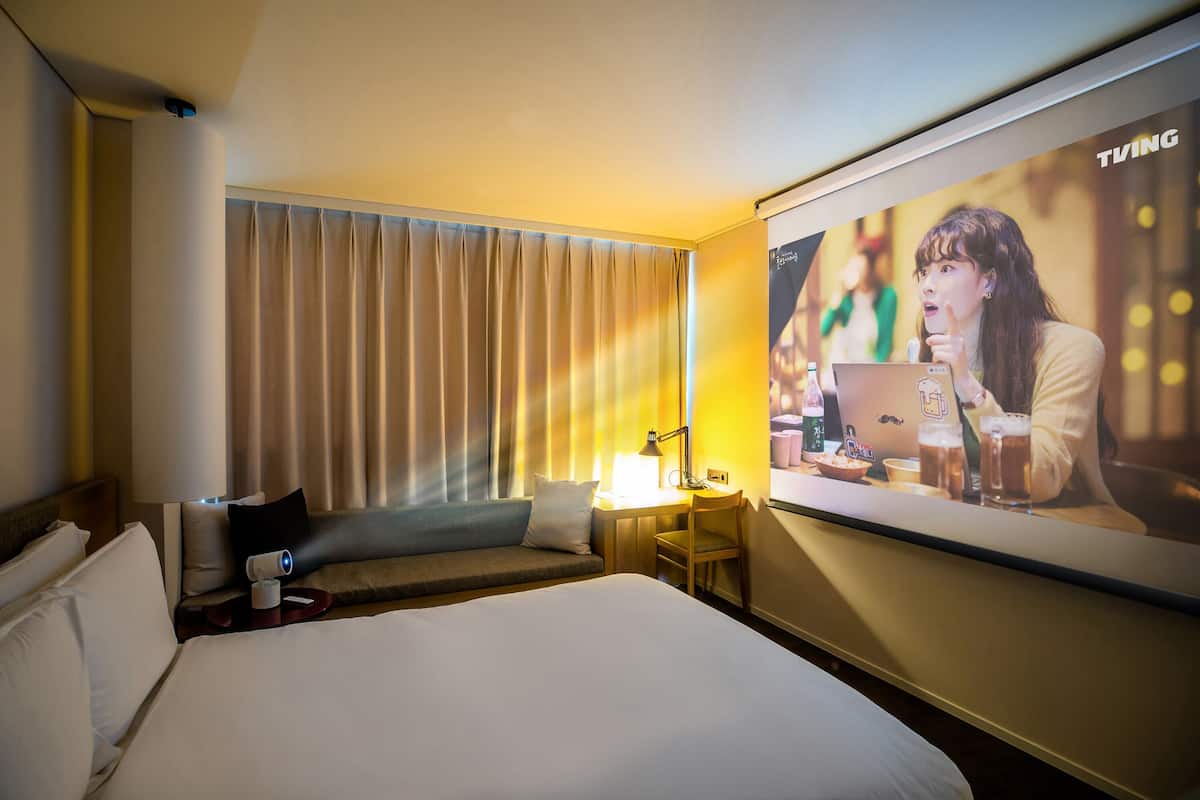 Hotellihuone, jossa on suuri sänky, työpöytä kannettavalla tietokoneella, tuoli, lamppu ja televisio, jolta näkyy jokin ohjelma.