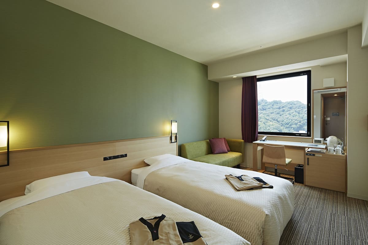 Ein Hotelzimmer mit zwei Betten, einem Schreibtisch, einem Stuhl, einem Fenster mit Blick und einer Minibar.