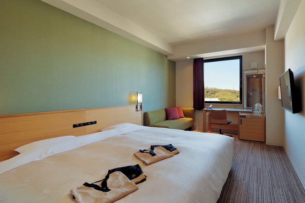 Ein Hotelzimmer mit einem großen Bett, einem Schreibtisch mit Stuhl, einem Fenster mit Aussicht und einem an der Wand befestigten Fernseher.