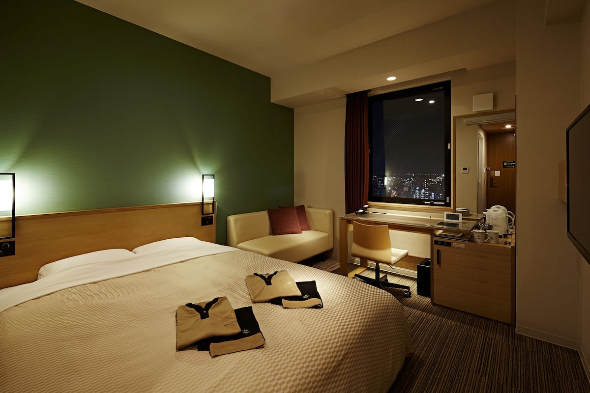 Ein Hotelzimmer mit einem großen Bett, einem Schreibtisch, einem Sessel und einer Couch, mit Blick auf die beleuchtete Stadt.