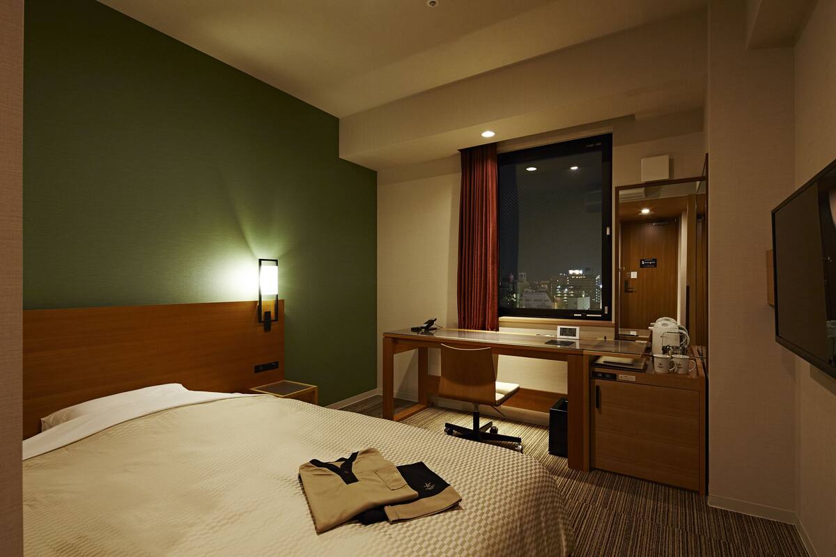 Ein Hotelzimmer mit Bett, Schreibtisch, Stuhl, Fernseher und Fenster mit Blick auf die Stadt.