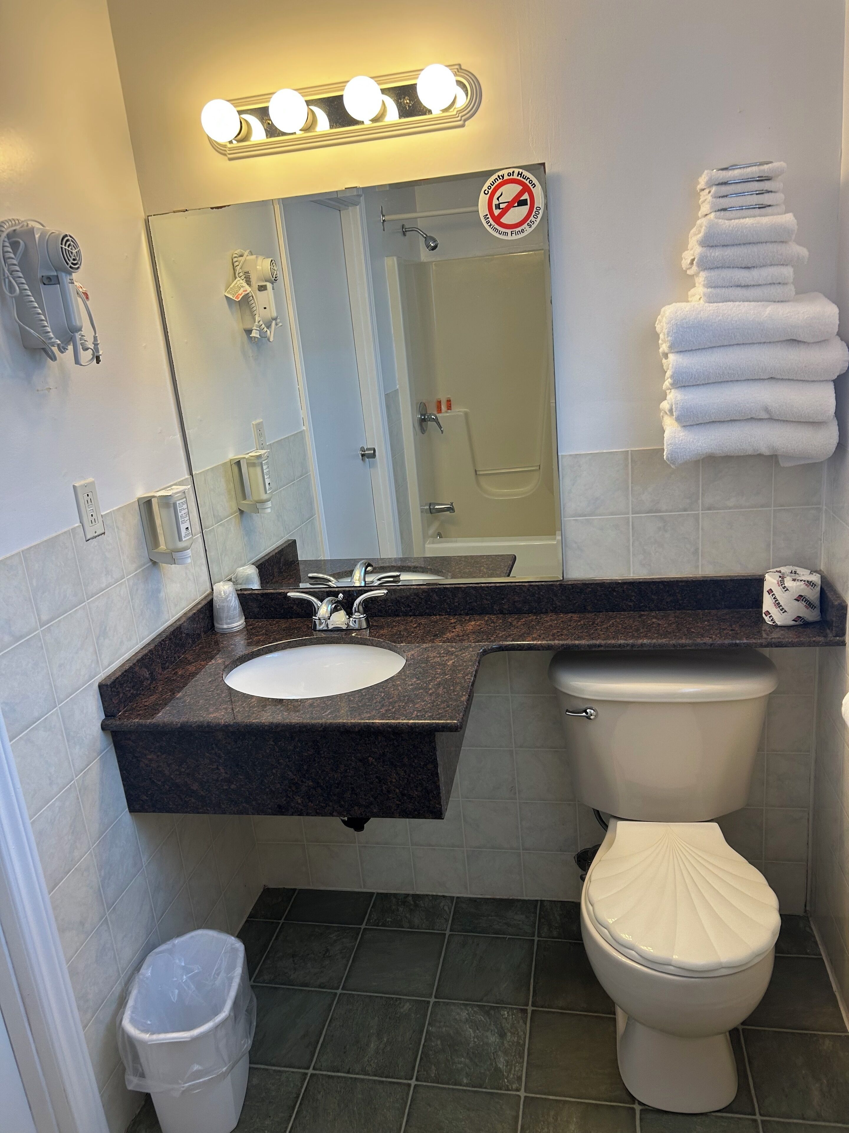 Ein Badezimmer mit Waschbecken, Toilette und Duschkabine. Handtücher sind an der Wand gestapelt und ein Mülleimer steht bereit.