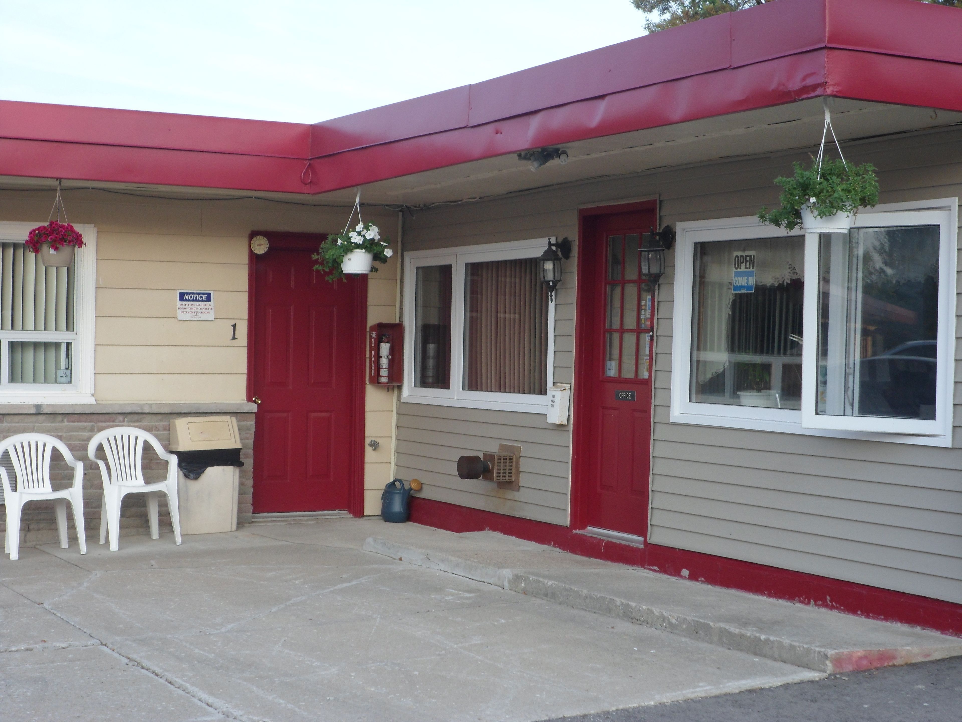 Un motel avec une porte rouge, un panneau d’affichage et deux chaises en plastique blanches devant l’entrée.
