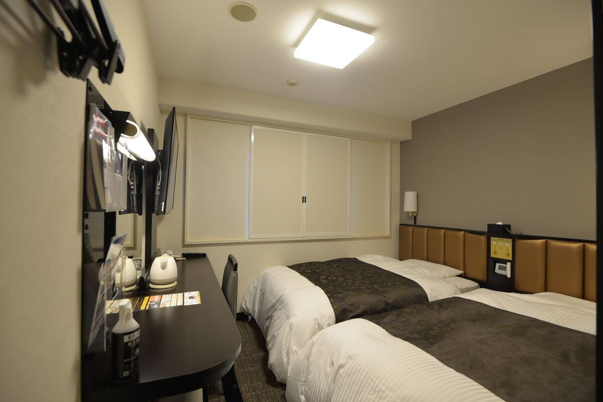 A hotel room with two beds, a desk with a telephone, a mirror, and a window with blinds.