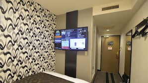 A hotel room with a bed, a TV mounted on the wall displaying a hotel directory, a patterned curtain, and a hallway leading to the door. - APA Hotel Nishimaizuru-Ekimae (Maizuru)