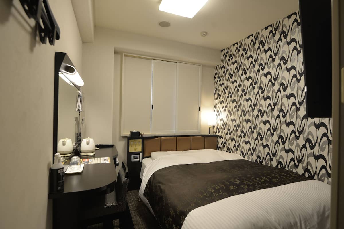 A hotel room with a bed, a desk with a lamp, a mirror, a nightstand, and a patterned curtain.