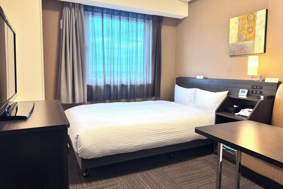 A hotel room with a large bed, a desk, a chair, a nightstand, a lamp, and a window with curtains.
