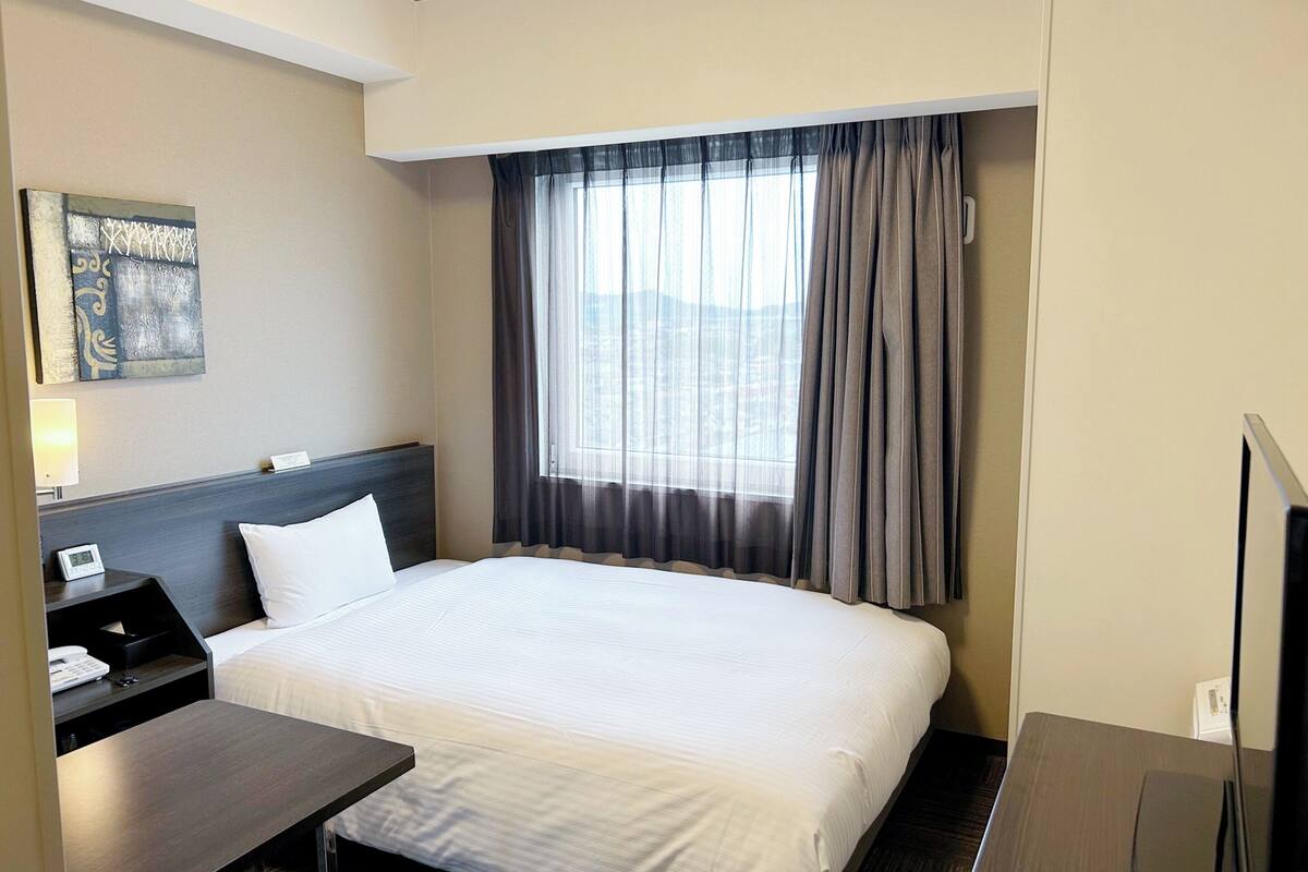 A hotel room with a bed, a desk, a chair, a TV, a window with curtains, and a painting on the wall.