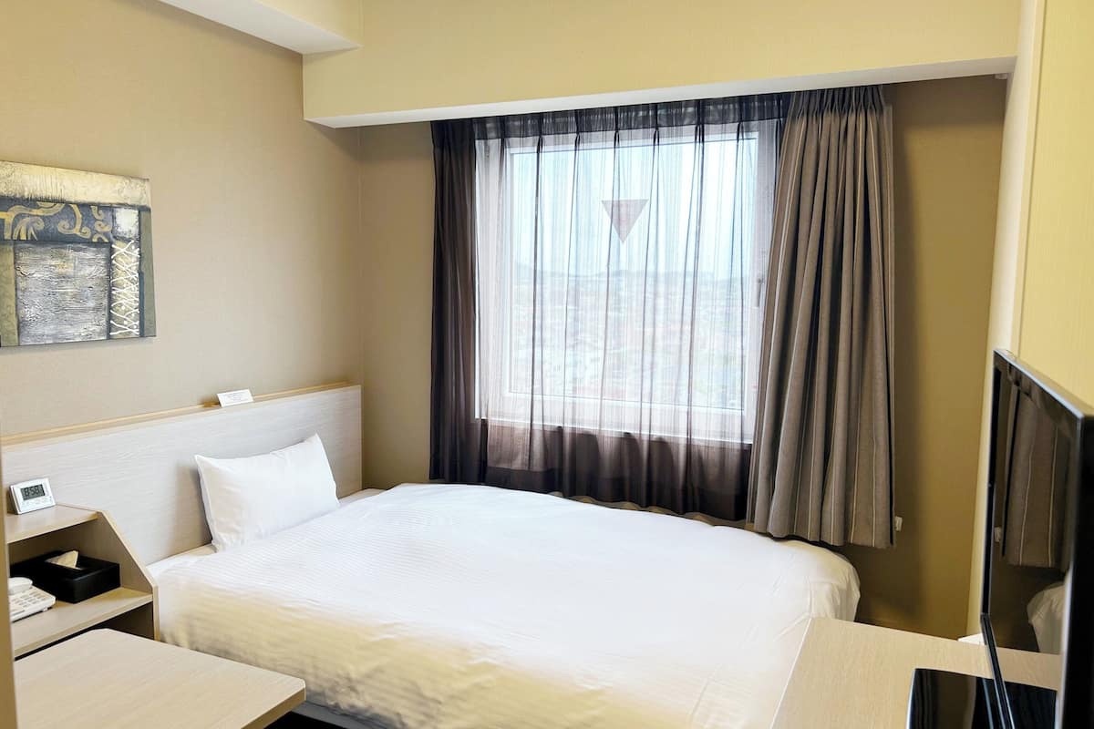 A hotel room with a bed, bedside table, and a window with curtains.