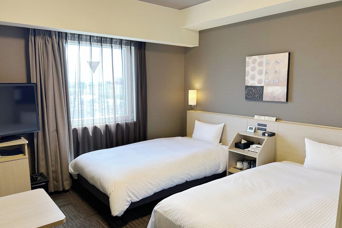 A hotel room with two beds, a TV, a bedside table, a lamp, and a painting on the wall.