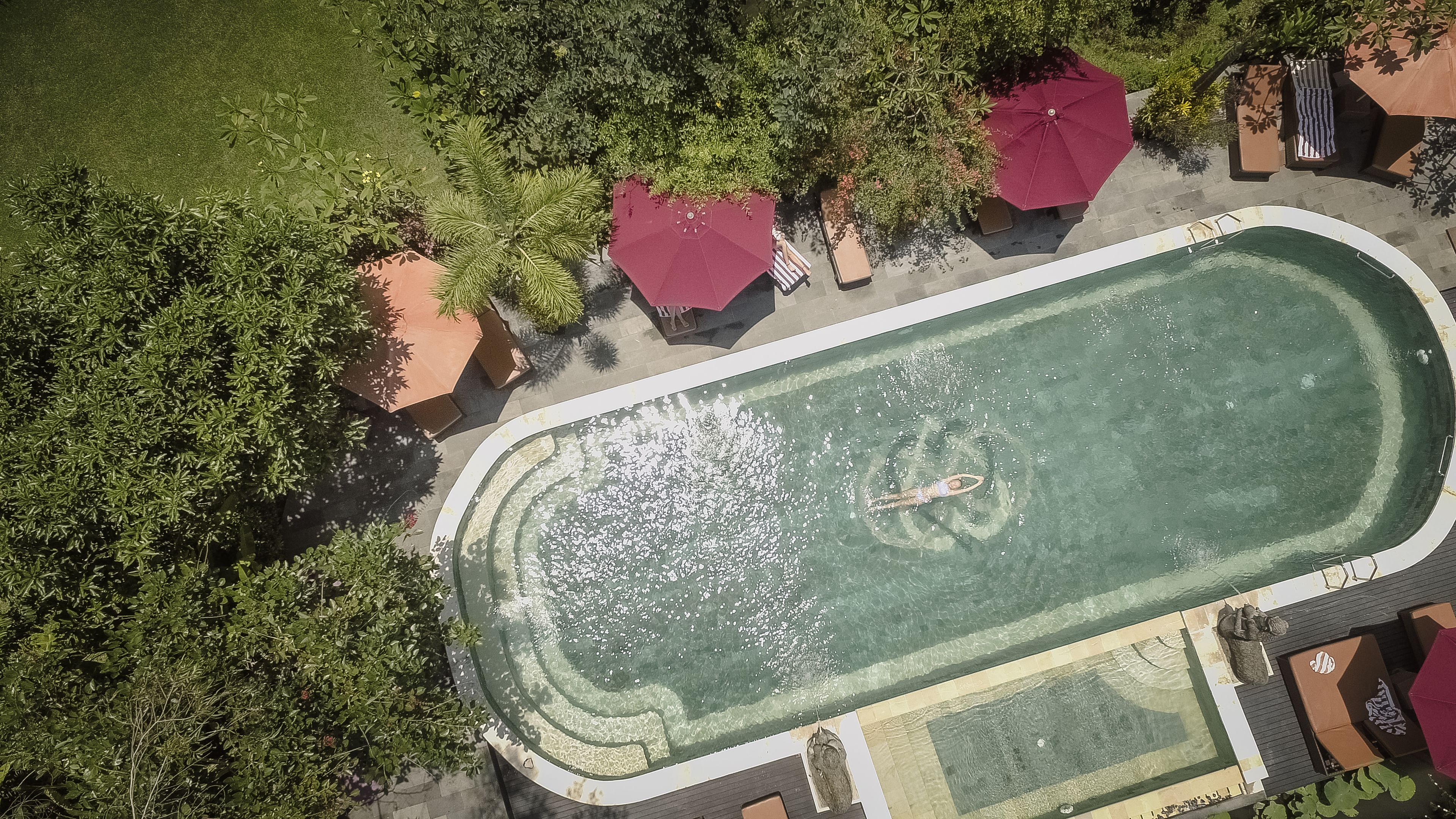 An aerial view of a swimming pool surrounded by lush greenery and shaded by red umbrellas.