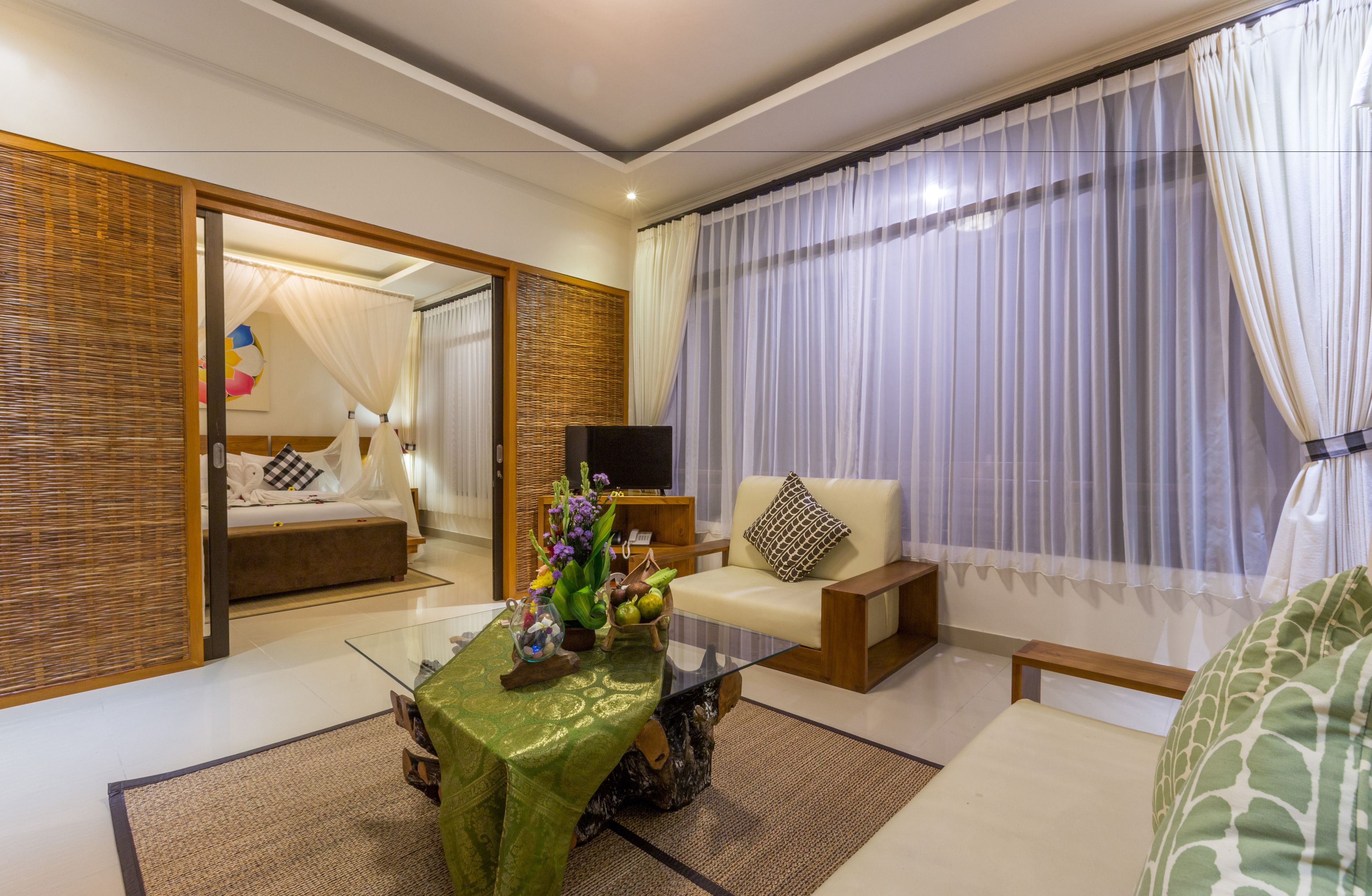 A modern hotel room with a glass coffee table, a sofa, a bed with a canopy, and a large window with sheer curtains.