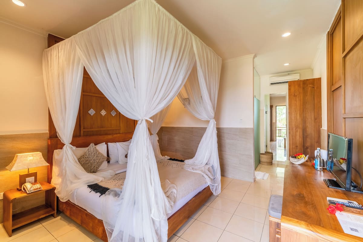 A bedroom with a four-poster bed, a bedside table with a lamp, a TV, and a wooden desk with a computer and some papers.
