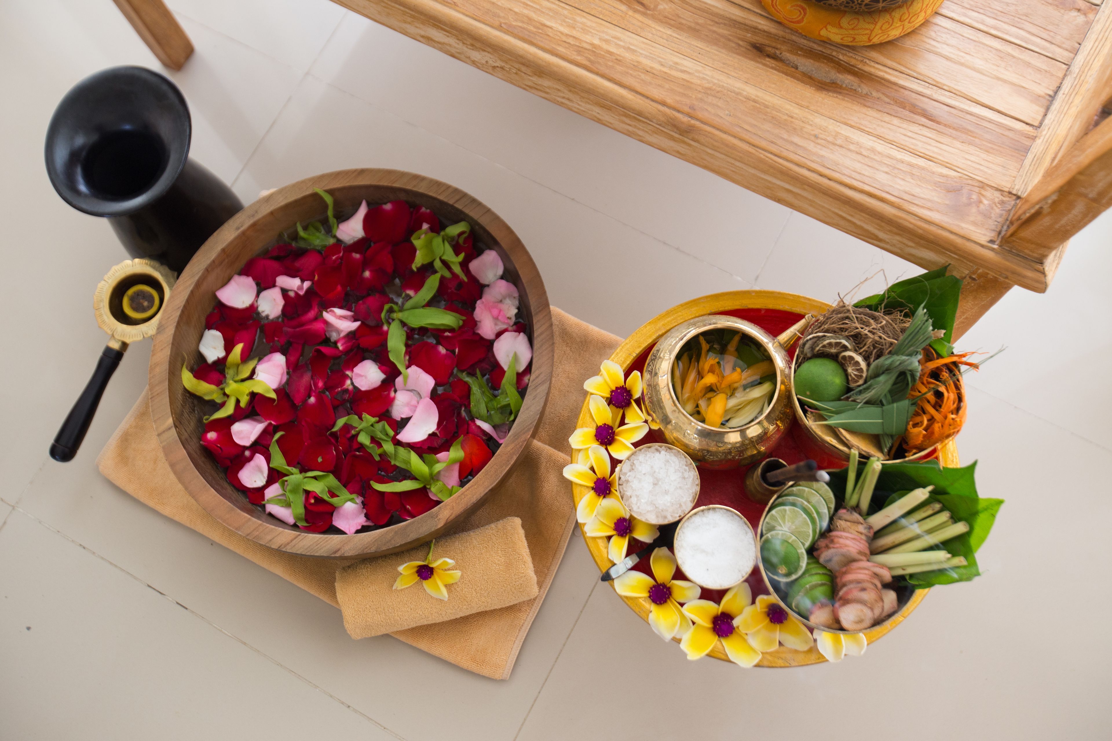 A wooden table with a bowl of rose petals, a tray with various herbs and spices, and a black container.