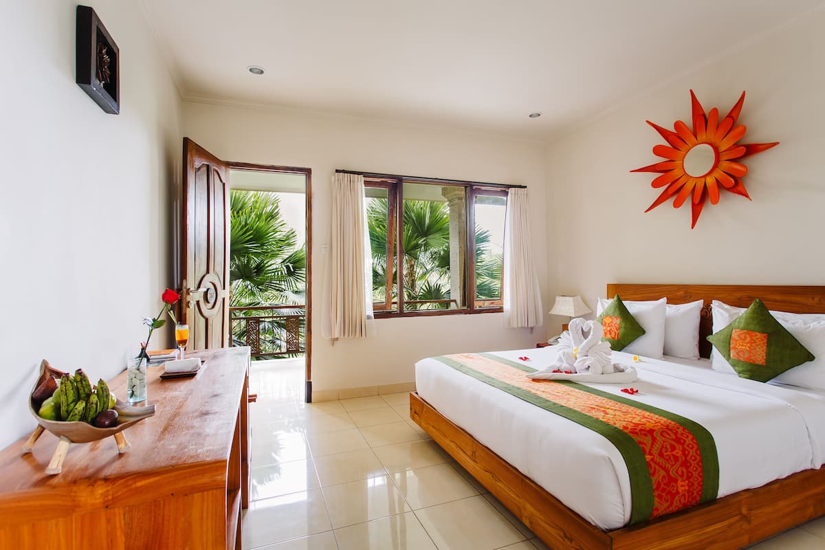 A hotel room with a bed, a desk with a fruit bowl, a window with curtains, and a decorative sun on the wall.