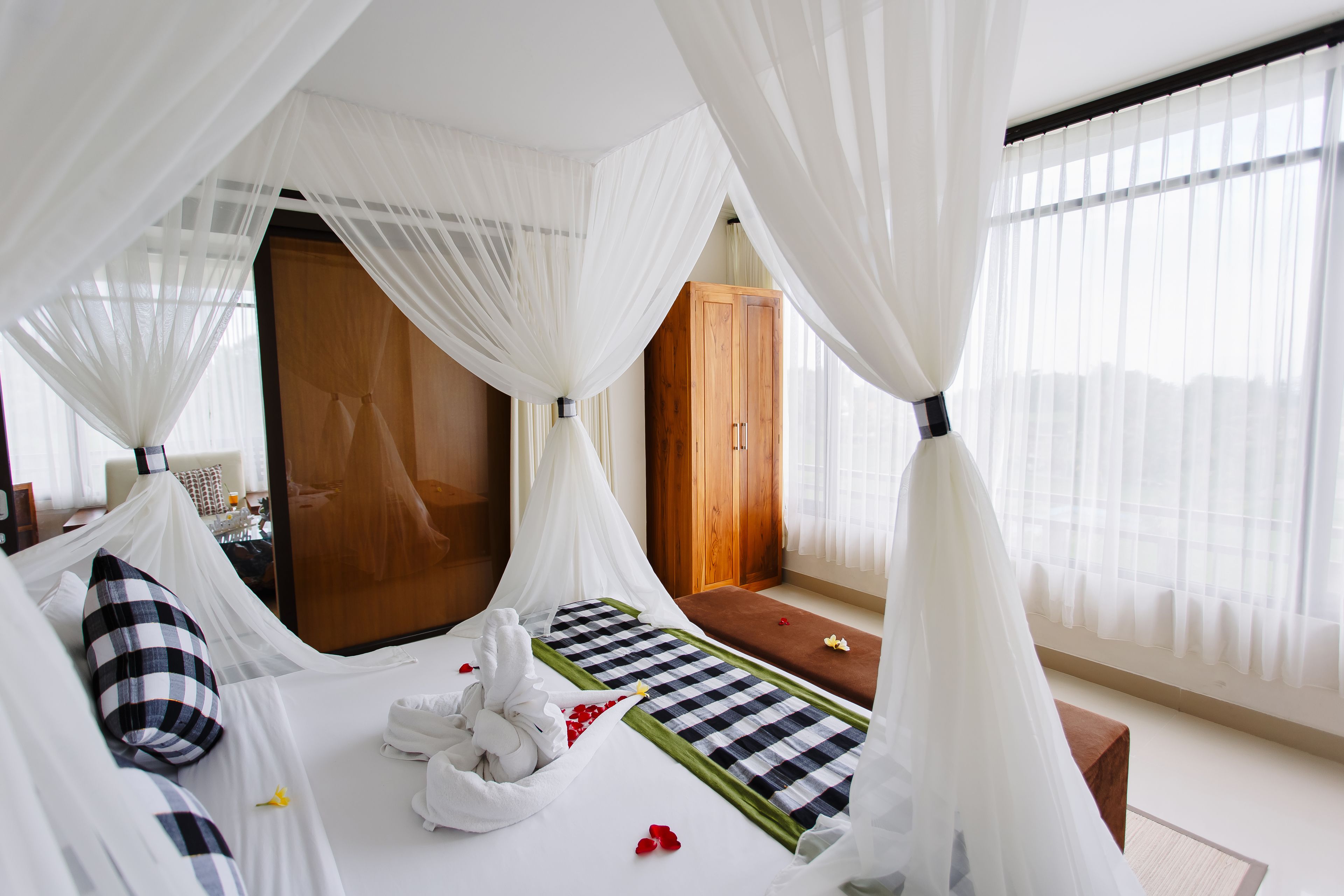 A hotel room with a canopy bed, a wooden wardrobe, and a view of the city through sheer curtains.