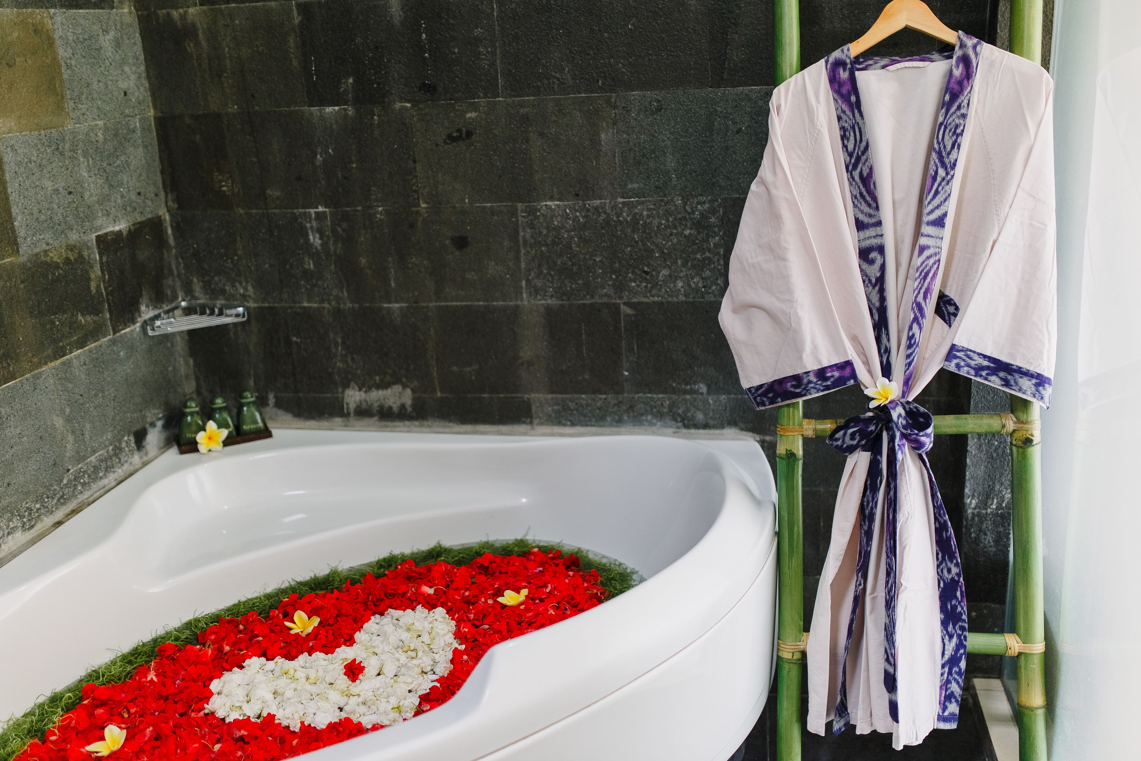 A bathtub filled with red and white flowers, a white robe tied to a bamboo stand, and a stone wall in the background.