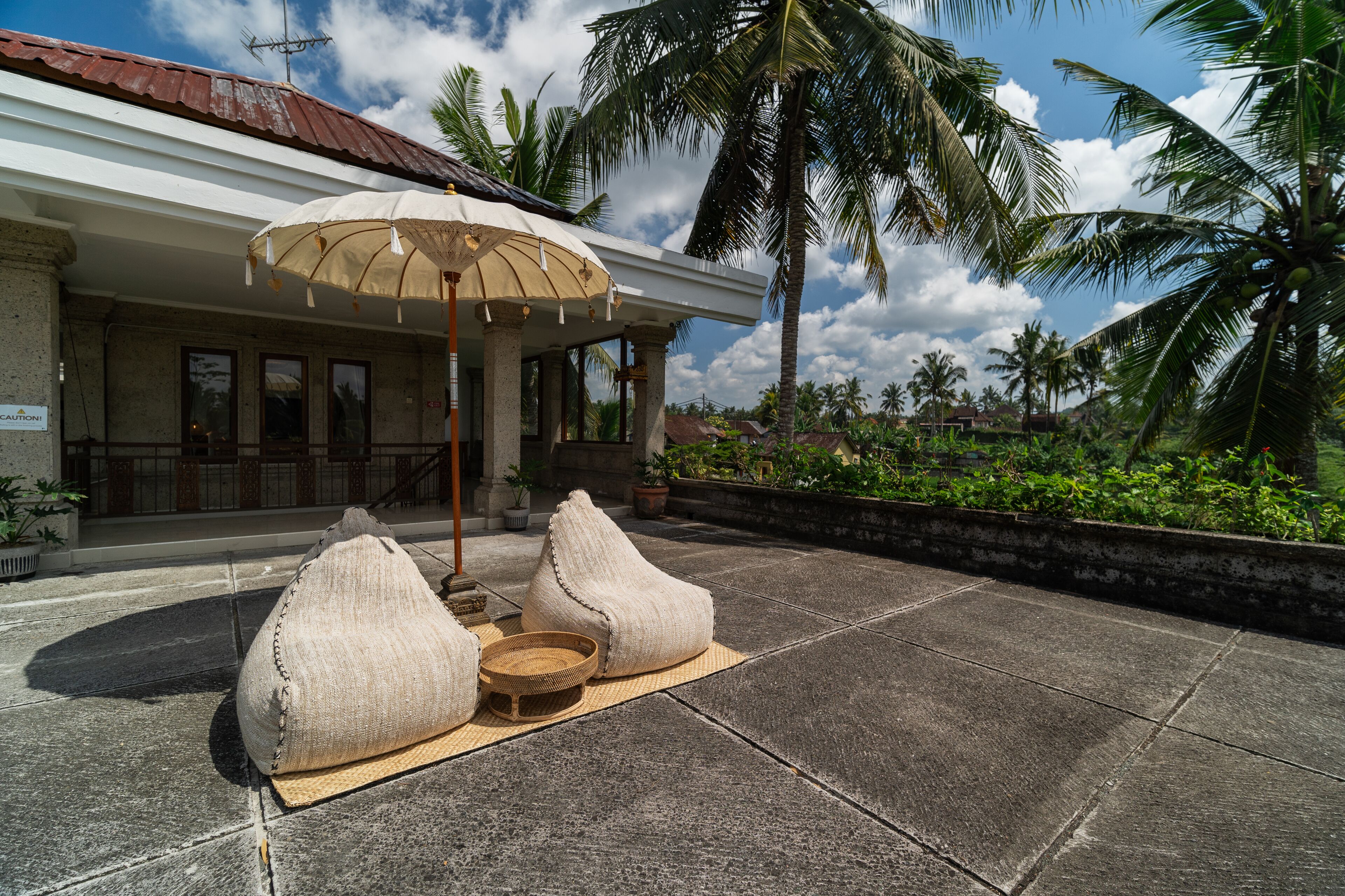 A covered outdoor seating area with a table and two bean bags, a tiled patio, and a building with a red roof and a sign that reads "CAUTION".