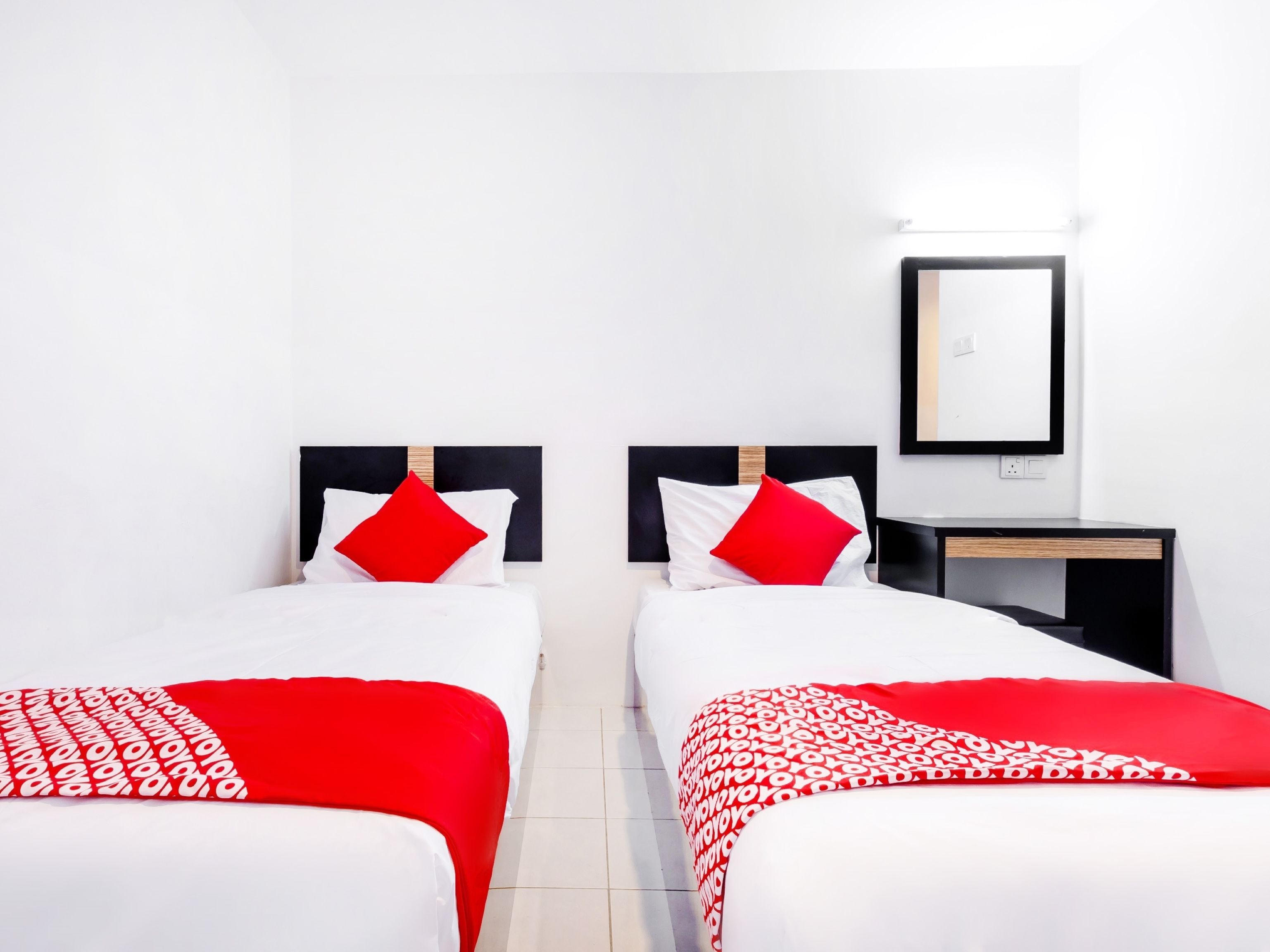 Ein Hotelzimmer mit zwei Betten, jeweils mit weißer Bettwäsche und roten Akzenten, einem kleinen Nachttisch und einem an der Wand befestigten Spiegel.