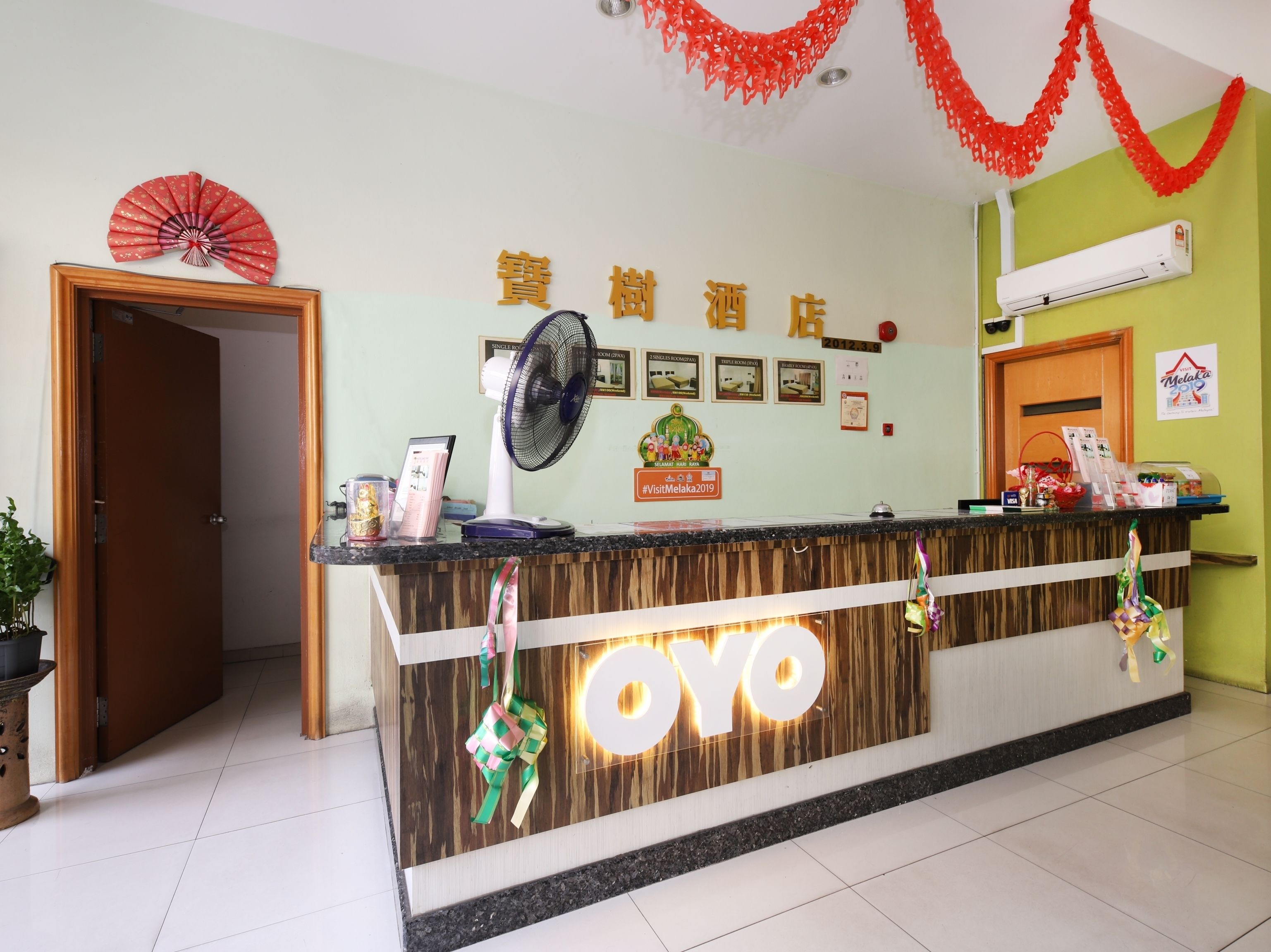 A hotel reception desk with a wooden finish and a prominent 'OYO' logo. There is a fan on the wall, a framed certificate, and a red decorative item hanging above the reception.