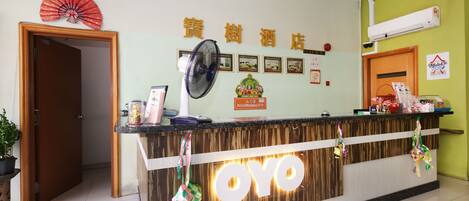 A hotel reception desk with a wooden finish and a prominent 'OYO' logo. There is a fan on the wall, a framed certificate, and a red decorative item hanging above the reception.