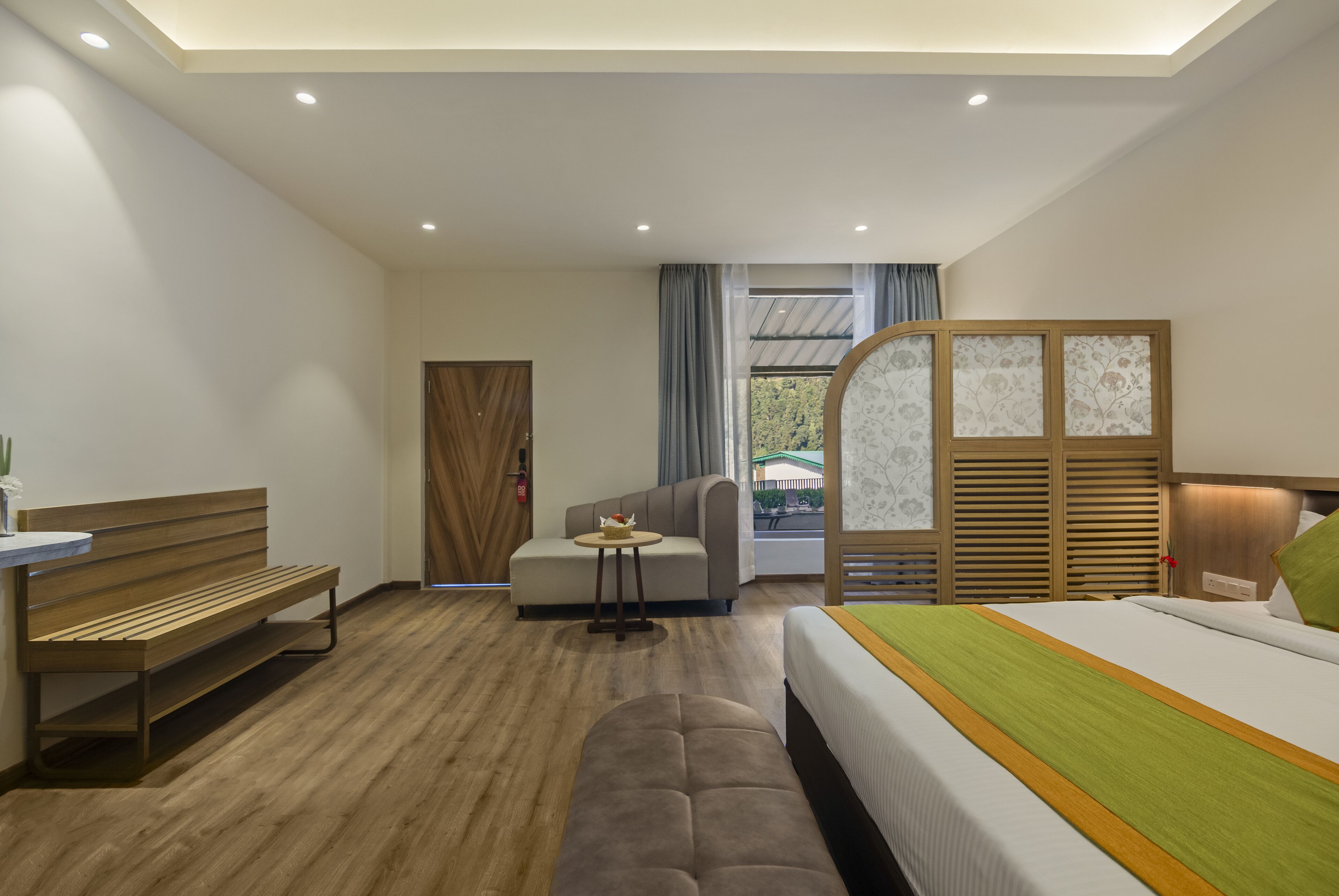 A modern hotel room with a wooden bench, a sofa, a bed with a green and yellow bedspread, and a view of the outdoors through a window with curtains.