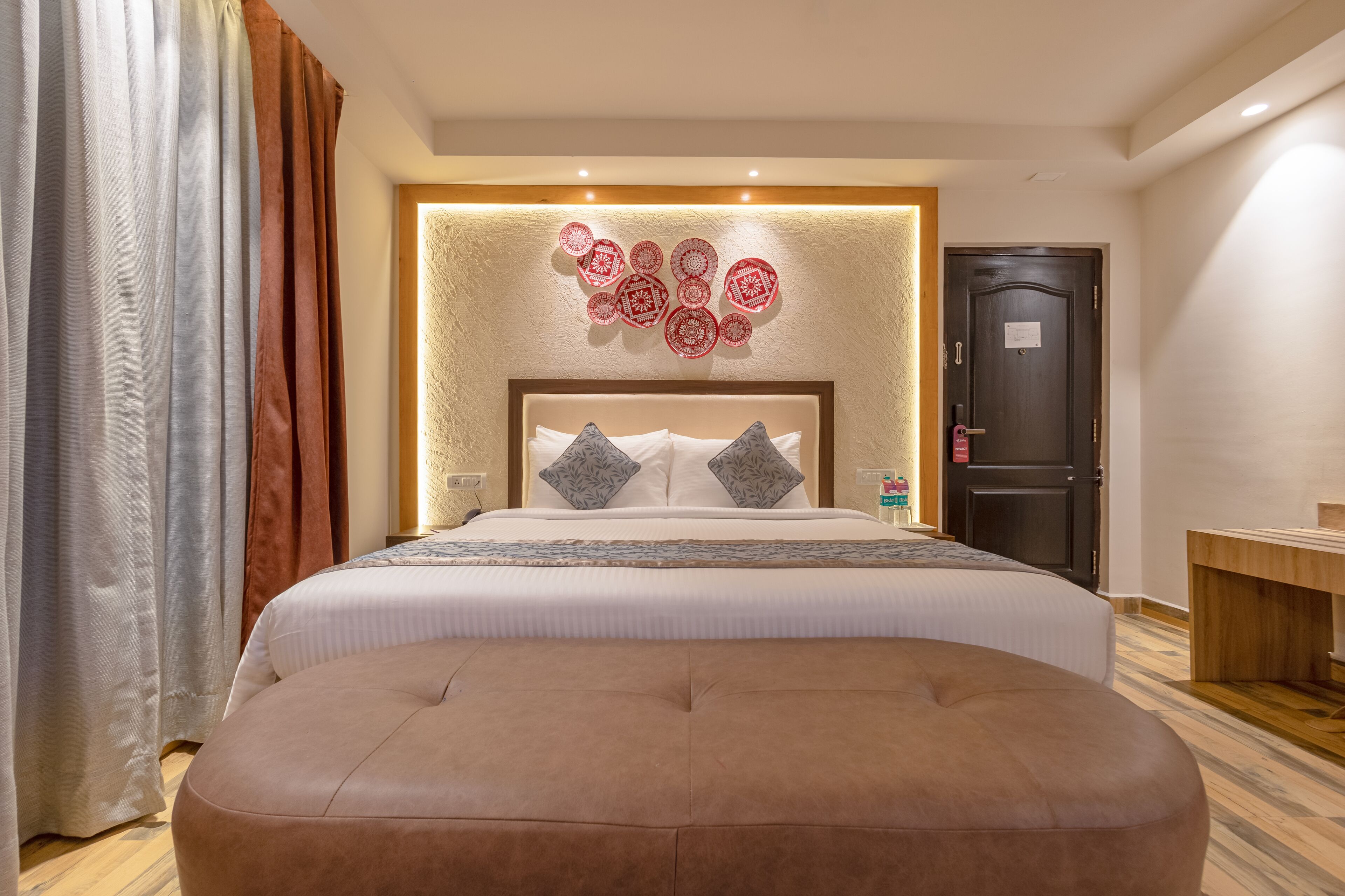 Habitación de hotel con una cama grande, cabecera de madera, panel decorativo en la pared, mesita de noche con una botella y una puerta con un letrero.