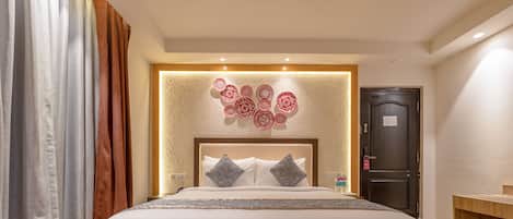 Une chambre d’hôtel avec un grand lit, une tête de lit en bois, un panneau décoratif fixé au mur, une table de chevet avec une bouteille et une porte avec une pancarte.