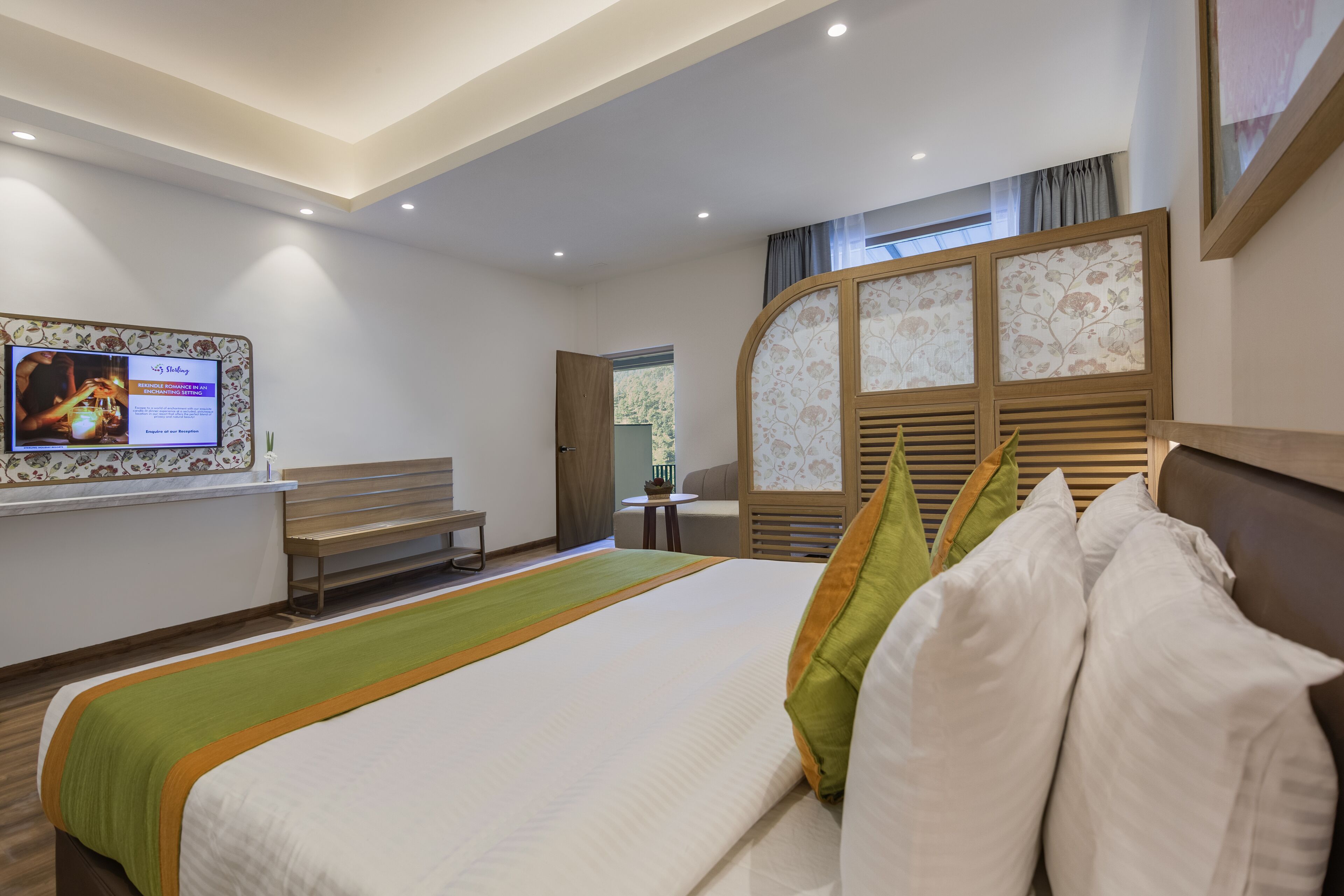 A hotel room with a large bed, a television mounted on the wall, a small table, and a window with curtains.