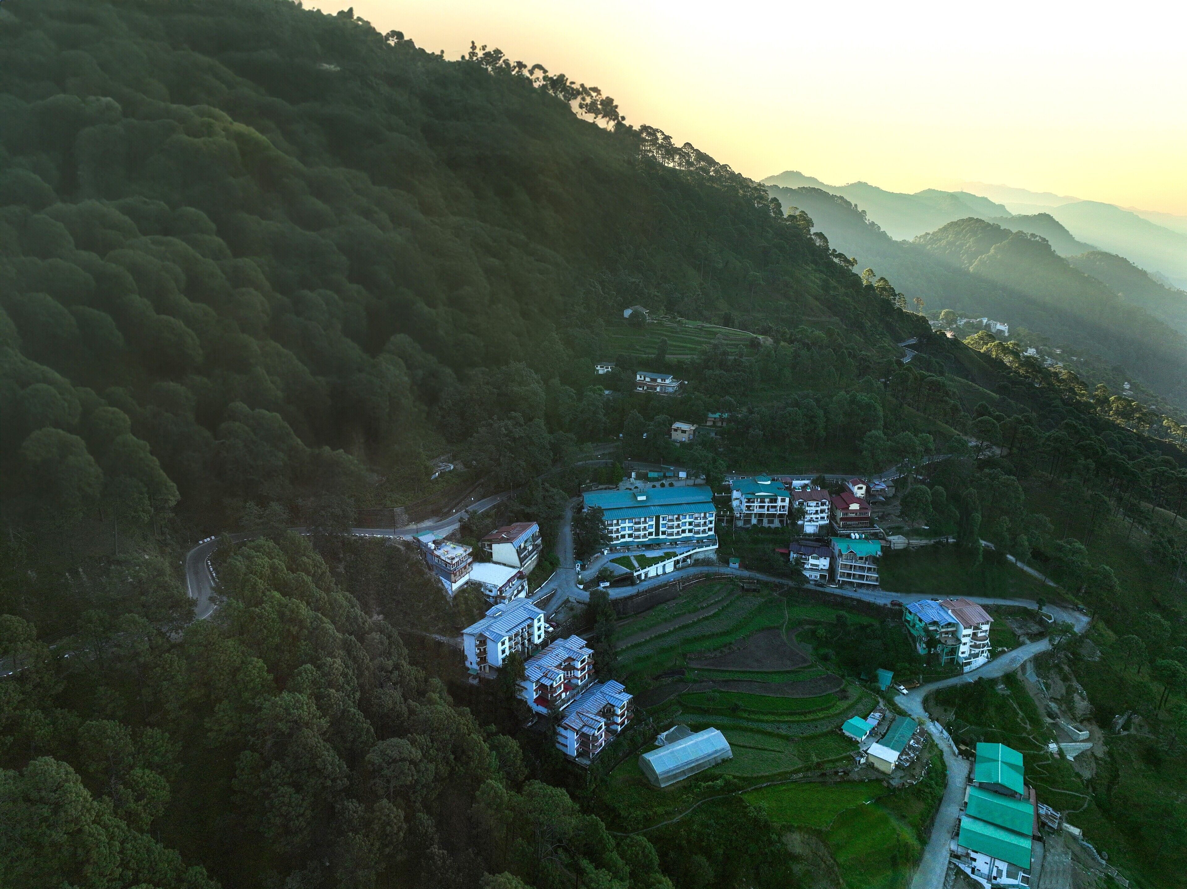 An aerial view of a rural village with terraced fields, a winding road, and several buildings with flat roofs.