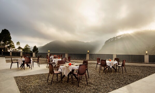 An outdoor dining area with tables and chairs, set against a backdrop of misty mountains and a cloudy sky.