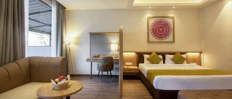 A hotel room with a sofa, a desk, a bed with two pillows, a small round table with a basket, and a large window with curtains.